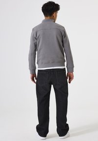 Hombre de pie de espaldas a la cámara, vistiendo una chaqueta gris, jeans negros, zapatillas blancas y una camisa blanca, contra un fondo blanco liso.
