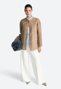 Beige wollen jas met een ronde halslijn en vier knopen aan de voorkant, gecombineerd met een lichtblauw gestreept overhemd en brede witte broek. Houdt een donker bontaccessoire vast.