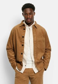 Braune Cordjacke mit spitzem Kragen, Knopfverschluss und Brusttasche, getragen über einem hellen Hemd mit Knöpfen.