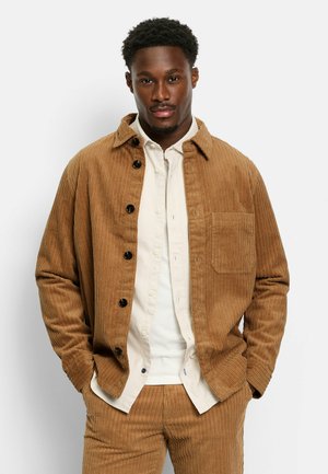 Braune Cordjacke mit spitzem Kragen, Knopfverschluss und Brusttasche, getragen über einem hellen Hemd mit Knöpfen.