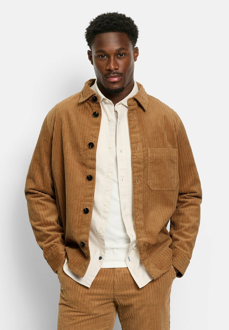 Braune Cordjacke mit spitzem Kragen, Knopfverschluss und Brusttasche, getragen über einem hellen Hemd mit Knöpfen.