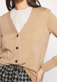 Beige stickad cardigan med V-ringning, försedd med fyra mörka knappar och ribbad textur. Bärs över en ljus skjorta och mönstrad kjol.