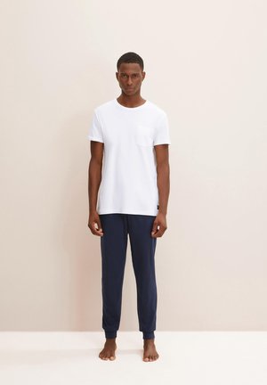T-shirt blanc à manches courtes avec une poche associé �à un pantalon de jogging bleu marine, tous deux en tissu doux et lisse. Le modèle se tient pieds nus contre un fond neutre.