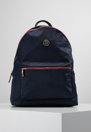 Granatowy plecak z przednią kieszenią na suwak, czerwonymi i złotymi akcentami zamka oraz okrągłym metalowym logo na górnym przednim panelu.