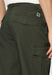 Cargo-Hosen aus dunkelgr�ünem Stoff mit strukturierter Oberfläche, ausgestattet mit Seitentaschen und einer Gesäßtasche. Enthält ein kleines Markenlogo-Patch.