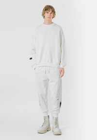 Sudadera blanca con cuello redondo y panel frontal decorativo, combinada con pantalones de chándal blancos que tienen acentos gráficos. Usada con botas claras.