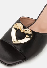 Black leather open-toe shoe with a glossy gold heart accent on the front. Features a smooth texture and a square toe design.