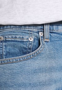 Světle modré džíny z denimu s texturovaným povrchem, vybavené stříbrným kováním, malou přední kapsou a kontrastním prošíváním podél švů.