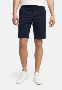 Shorts en coton bleu marine à coupe droite, avec poches avant et passants de ceinture, associés à des baskets blanches et un t-shirt blanc simple.