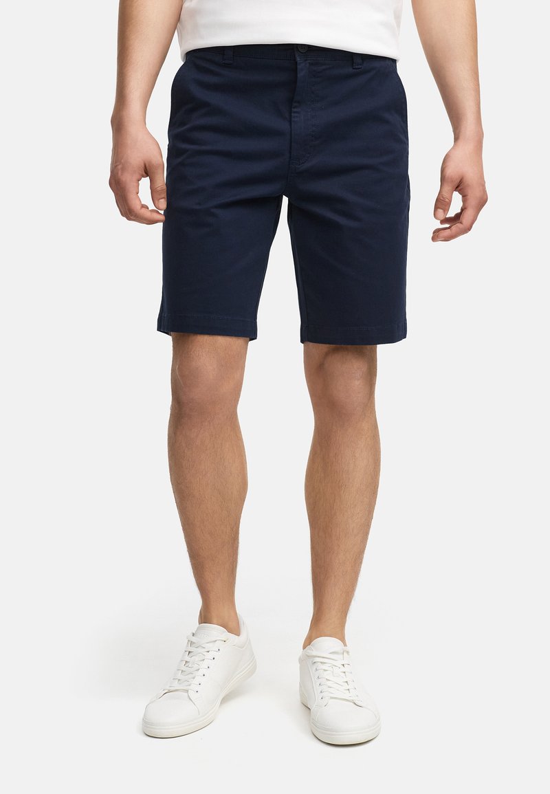 Shorts en coton bleu marine à coupe droite, avec poches avant et passants de ceinture, associés à des baskets blanches et un t-shirt blanc simple.