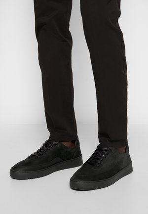 Person wearing black slim-fit pants and black low-top sneakers standing on a light gray floor against a white background.