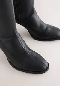 Sorte læderstøvler med spids tå, glat overflade, sidelås med lynlås, gummisål og kontrastsyning.