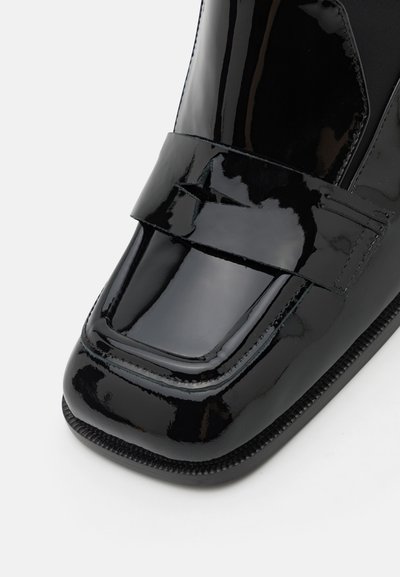 Chaussure en cuir verni noir avec un bout carré et une large sangle au milieu du pied avec des détails cousus. Fini lisse et brillant.