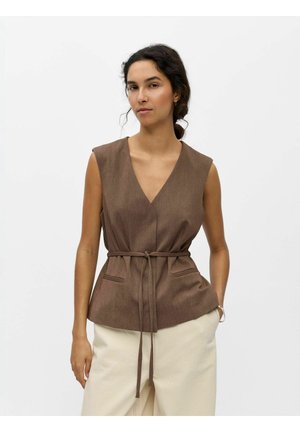 Donna con capelli scuri raccolti dietro, che indossa un gilet marrone senza maniche con scollo a V e cintura annodata in vita, e pantaloni beige su uno sfondo neutro.