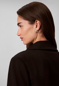 Femme aux longs cheveux bruns portant des boucles d'oreilles argentées en forme de créoles et un manteau noir, vue de profil sur un fond gris uni.