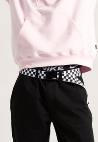 Pantaloni neri con una cintura a scacchi bianchi e nera, indossati con una felpa rosa chiaro con tasca a canguro.