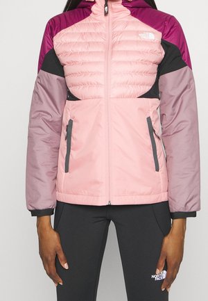 Femme portant une veste isolante North Face rose, violette et noire avec des poches zippées et des leggings noirs avec un logo sur la cuisse.