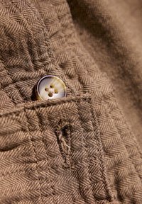 Tejido marrón texturizado con un patrón de espiga, que presenta un botón de color claro y un pequeño desgarro en la tela cerca del bolsillo.