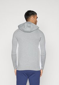 Gr�å hættetrøje med en tætsiddende design, der har hvide sidepaneler, en snoretrækshætte og tekstureret stof. Bagsiden har en rundet kant.