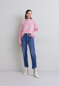 Maglione lavorato a maglia di colore rosa chiaro con un design a gradient, abbinato a jeans blu a vita alta e stivaletti alla caviglia bianchi con stampa pitone. Sfondo semplice.