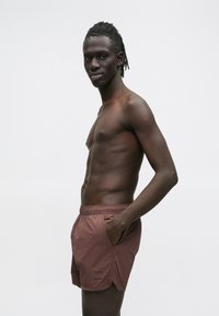 Bordowe spodenki kąpielowe z elastycznym pasem, bocznymi kieszeniami i lekko falowanym dołem. Wykonane z lekkiego, gładkiego materiału.