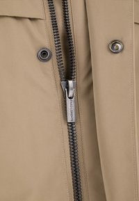 Beżowa kurtka z widocznym zamkiem błyskawicznym i zatrzaskiem. Zamek posiada metalowy uchwyt z napisem "Pako Lorente." Gładka tekstura materiału.