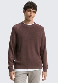 Bruine gebreide trui met ribbeltextuur, ronde hals, en lange mouwen, gelaagd over een wit shirt, gedragen met lichtgekleurde broek.