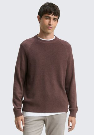 Brauner Strickpullover mit geripptem Muster, Rundhalsausschnitt und langen Ärmeln, über einem weißen Hemd getragen, kombiniert mit hellen Hosen.