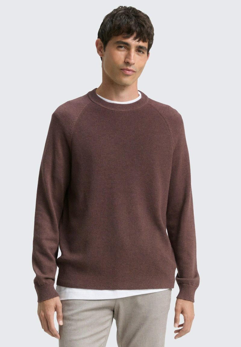 Bruine gebreide trui met ribbeltextuur, ronde hals, en lange mouwen, gelaagd over een wit shirt, gedragen met lichtgekleurde broek.