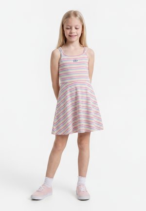 Μικρό κορίτσι στέκεται χαμογελώντας, φοράει αμάνικο πολύχρωμο ριγέ φόρεμα με το κείμενο "GAP", λευκές κάλτσες και ροζ αθλητικά παπούτσια σε λευκό φόντο.