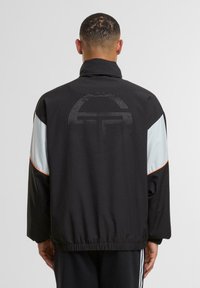 Juodas švarkas su šviesiai mėlynomis rankovėmis ir oranžiniais akcentais. Nugarėlėje - taškinis logotipas, sudarytas iš apskrito rašto. Elastinga talija ir rankogaliai.