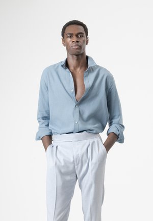 Hombre con cabello corto y rizado que lleva una camisa azul claro desabrochada y pantalones blancos plisados, de pie con las manos en los bolsillos.