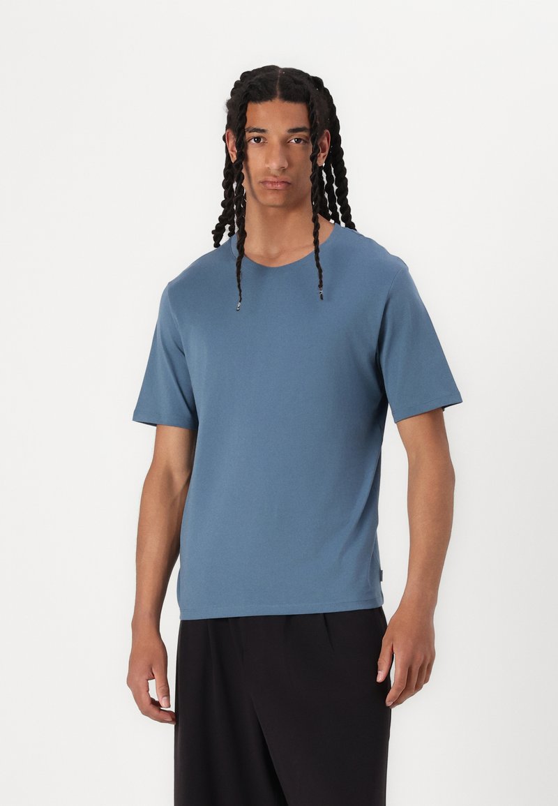 Blue cotton T-shirt featuring a round neckline and short sleeves, worn with black loose-fitting trousers. Smooth texture, minimal design.