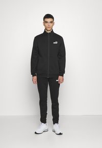 Puma tracksuit in solid black, featuring a zip-up jacket and matching pants with logo details. Material appears soft and textured. White sneakers.