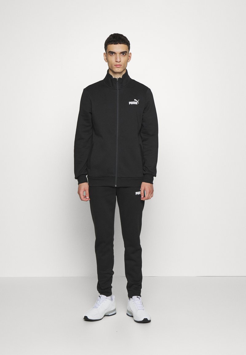 Puma tracksuit in solid black, featuring a zip-up jacket and matching pants with logo details. Material appears soft and textured. White sneakers.