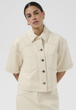 Femme aux cheveux foncés portant des boucles d’oreilles créoles dorées et une veste crème boutonnée à manches courtes avec poches avant, sur un fond uni.