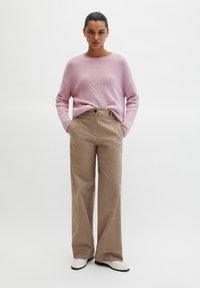 Femme debout les mains dans les poches, portant un pull côtelé rose, un pantalon beige à jambes larges et des chaussures blanches, sur un fond blanc.