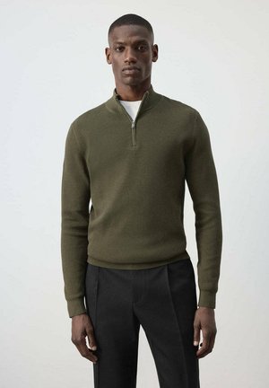 Man staat met een olijfgroene gebreide ritstrui over een wit hemd en zwarte broek tegen een effen lichte achtergrond.