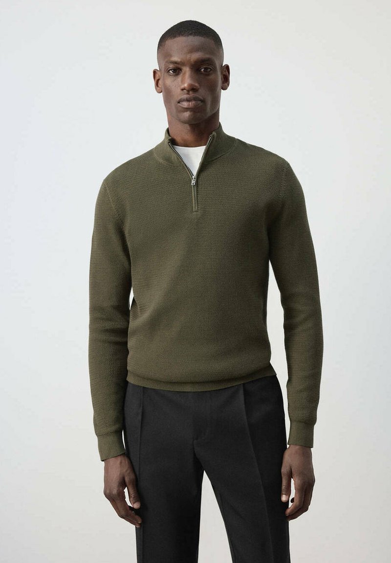Homme debout portant un pull en maille zippé vert olive sur une chemise blanche et un pantalon noir, sur un fond clair uni.