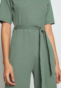 Ribbed jumpsuit in saliegroen, met een tailleband. De stof heeft een zachte textuur met korte mouwen en een aansluitende silhouet.