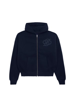 Mand med kort hår og øreringe iført en mørkeblå hættetrøje med lynlås og mørke jeans, står mod en ensfarvet lys baggrund.