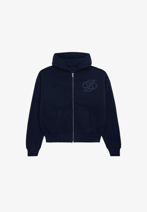 Mand med kort hår og øreringe iført en mørkeblå hættetrøje med lynlås og mørke jeans, står mod en ensfarvet lys baggrund.