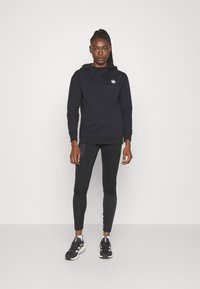 Haut à capuche noir avec cordon de serrage et un petit emblème, associé à des leggings noirs et des chaussures de sport. Le tissu semble lisse et extensible.