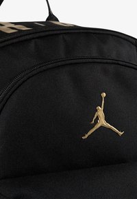 Rucsac din țesătură neagră, cu un logo Jumpman auriu proeminent, suprafață texturată și închidere cu fermoar. Accentele includ un nume de marcă îndrăzneț.