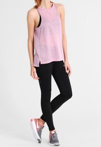 Top de malla rosa con aberturas laterales, combinado con leggings negros. El modelo lleva zapatillas deportivas en gris y coral, que cuentan con una superficie texturizada.