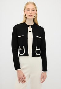 Cardigan nero corto con rifiniture bianche, due tasche frontali, dettagli dorati e un'apertura a chiave. Tessuto morbido e design aderente.