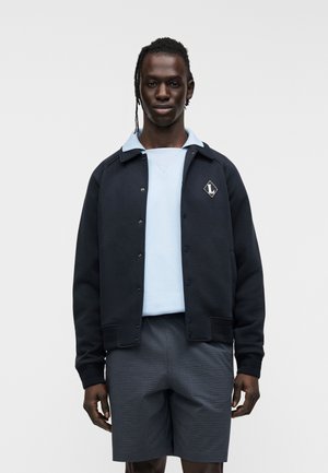 Granatowa kurtka na zamek z jasnoniebieskim kołnierzem, czarną naszywką z logo, w zestawieniu z jasnoniebieską bluzą z okrągłym dekoltem i szarymi, kraciastymi szortami.