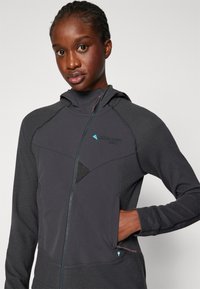 Schwarze Zip-Jacke aus strukturiertem Stoff mit Kapuze, geometrischer Panelstruktur und einem dezenten Logo auf der Brust.
