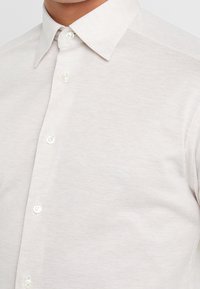 Camicia button-up color beige chiaro con tessuto strutturato, colletto classico e sei bottoni perlati lungo la pattina frontale.