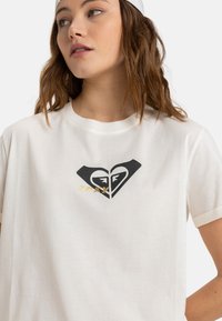 Biely bavlnený tričko s čiernym logom srdca a dizajnom tváre, a zlatým textom "roxy". Krátke rukávy a voľný strih.